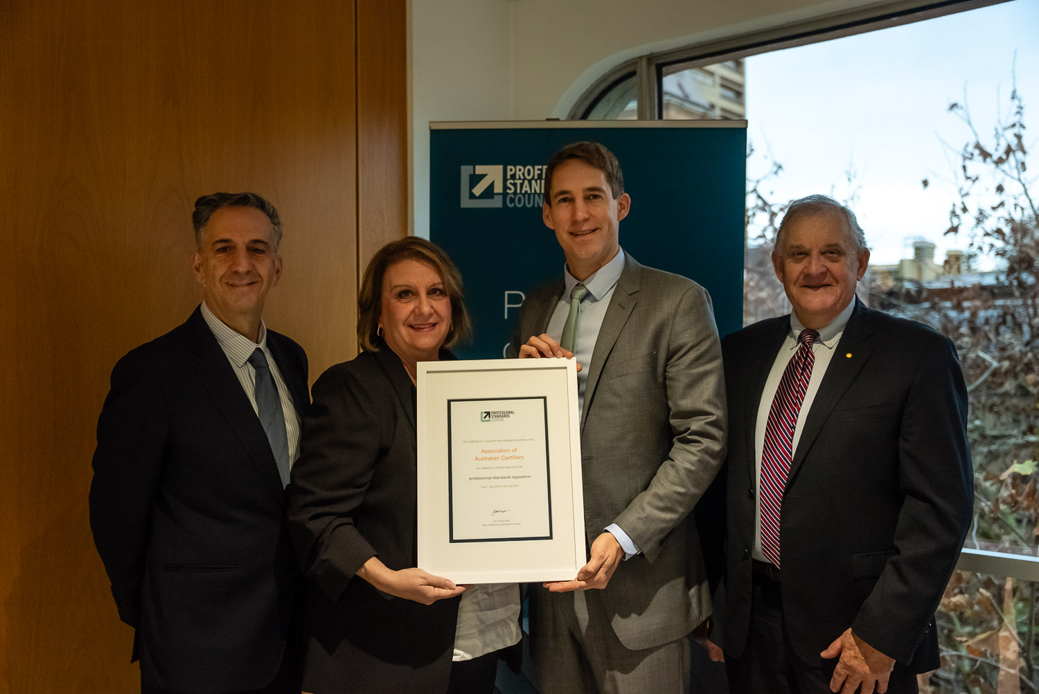 Photograph of four individuals standing indoors, with two holding a framed certificate. (L-R) Robert Marinelli (President, AAC), Jill Brookfield (CEO, AAC), Andreas Heger (CEO, PSC) and John Vines OAM (Chair, PSC) at the certificate presentation
